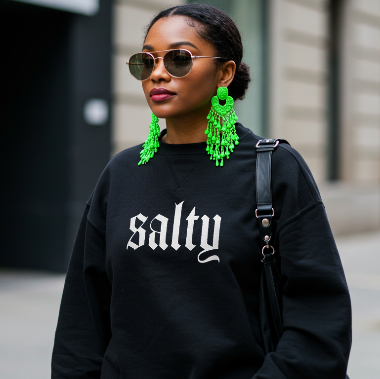 Woman wearing a black sweatshirt with 'salty' text, green earrings, and sunglasses on a city street.