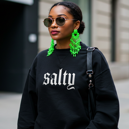 Woman wearing a black sweatshirt with 'salty' text, green earrings, and sunglasses on a city street.
